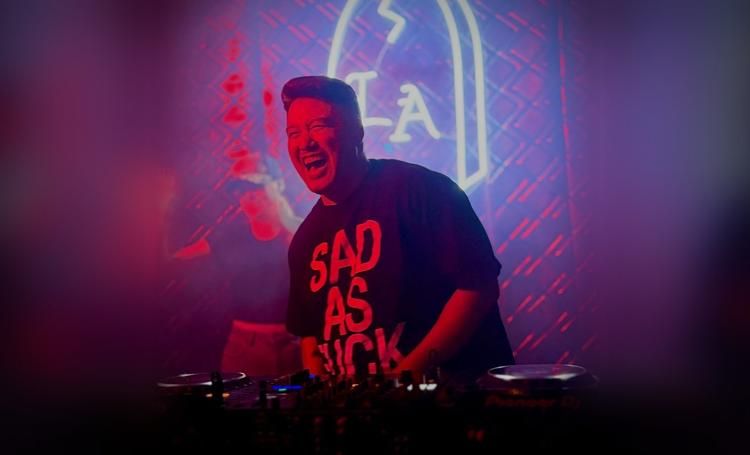 Chef Isaiah Liu laughing behind DJ decks under a neon sign